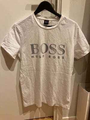 Vit t-shirt från Hugo Boss - Snygg vit t-shirt från Hugo Boss med stort BOSS-tryck på bröstet. Klassisk rund hals och korta ärmar. Tillverkad i mjuk bomull med regular fit för skön passform. Perfekt till jeans eller shorts för en clean och stilren look.