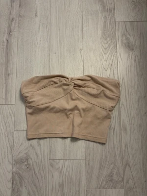 Beige ribbad bandeau tubtopp - Säljer en beige ribbad bandeau tubtopp med twistad detalj framtill. Toppen är axelbandslös och har en mjuk, stretchig passform som sitter snyggt på kroppen. Perfekt att styla till jeans eller kjol för en trendig look.