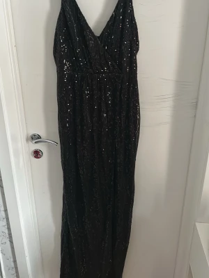 Svart paljettklänning med slits - Säljer en svart långklänning från Henriette med glittriga paljetter och en snygg slits på sidan. Klänningen har omlott upptill med v-ringning och smala axelband. Perfekt för dig som vill sticka ut på bal eller fest!