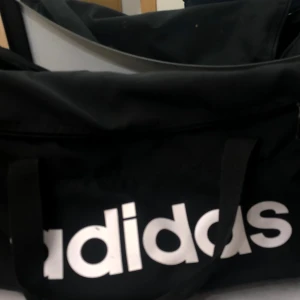Svart träningsväska från Adidas - Säljer en svart träningsväska från Adidas med stort vitt logotryck på sidan. Väskan har både handtag och justerbar axelrem, perfekt för gymmet eller sportaktiviteter. Tillverkad i slitstark polyester och har dragkedja upptill.