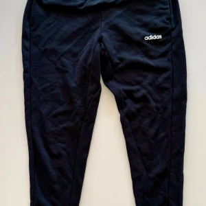 Svarta mjukisbyxor från Adidas - Svarta mjukisbyxor från Adidas med vit logga på låret. Byxorna har en smal passform och är tillverkade i mjukt bomullsmaterial. Perfekta för chill eller träning, med elastisk midja och avsmalnande ben.