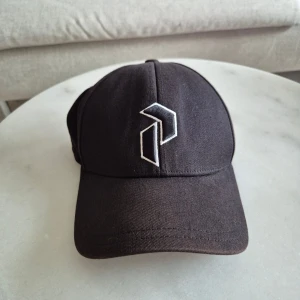Svart keps från Peak Performance - Svart keps från Peak Performance med vit broderad logga framtill. Klassisk böjd skärm. Tillverkad i bomull för en bekväm känsla. Perfekt accessoar för en sportig och stilren look. Storlek XL.