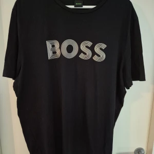 Svart BOSS t-shirt med tryck - Svart t-shirt från BOSS med stort vitt grafiskt BOSS-tryck på bröstet. Klassisk rund hals och korta ärmar. Tillverkad i mjuk bomull som känns skön mot huden. Perfekt för en clean och stilren look.