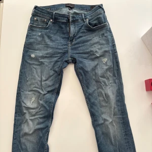 Snygga Junk De Luxe Jeans i bra skick -  Jeans från Junk de Luxe – Slim Fit Snygga blå jeans från märket Junk de Luxe. Modellen är Superflex Slim Fit, vilket innebär en smal passform med stretch för extra komfort. Jeansen har en tvättad look med slitningar framtill som ger en trendig och avslappnad stil.