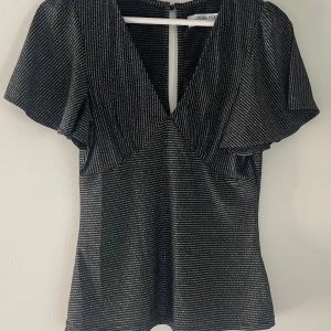 Glittrig fest topp - Snygg svart topp med silvriga glittriga ränder från Chiara Forti. Djup v-ringning framtill, korta vida ärmar och en liten knappdetalj i ryggen. Perfekt till fest och nyår!!