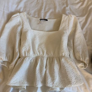 Vit blus med puffärm från Gina Tricot - Supersöt vit blus från Gina Tricot med fyrkantig ringning och puffiga trekvartsärmar. Blusen har en strukturerad, lite bubblig yta och volang nedtill som ger en romantisk vibe. Perfekt att styla med jeans eller kjol för en trendig look.