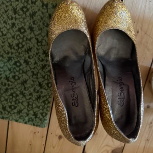 Glittriga guldiga pumps från Sit Sergio - Snygga pumps i glittrigt guld från Sit Sergio. Skorna har spetsig tå och hög klack, perfekta för att sticka ut. Ytan är täckt av glitter och insidan är i mörkare material. Riktigt statement-piece för dig som gillar att glänsa.