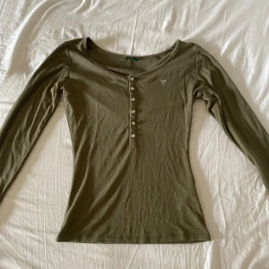 Grön henley/farfarströja från Guess - Olivgrön farfarströja från Guess. Har tappat några stenar i loggan. Strl XS/S, fraktar gärna tillsammans med andra plagg.