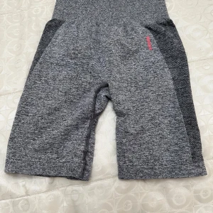 Grå träningstights shorts från Gymshark - Säljer ett par grå, högmidjade träningstights shorts från Gymshark med rosa logga bak i midjan. De är tillverkade i ett stretchigt och mjukt syntetmaterial, har en tight passform och är perfekta för gym eller annan träning.