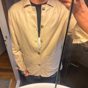 Beige overshirt - Stilren beige overshirt från Mango Man med klassisk krage, stora framfickor och knäppning med fyra knappar. Jackan är i ett lätt material och har en avslappnad passform, perfekt för lager-på-lager. Passar dig som gillar minimalistisk och clean stil. Nypris 1100kr