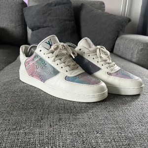 Louis Vuitton PRISM sneakers - Säljer nu mina Snygga Louis Vuitton sneakers i vitt skinn med pastellfärgade paneler i rosa, blått och lila. Skorna har klassiskt LV-monogram och vita snören. Perfekt för dig som vill sticka ut med exklusiva detaljer och unik färgkombination. Bortsett från jacket på ena skon(där av priset) är dom superfräscha. Skorna står mest på hyllan o är i behov av ny ägare .Lådan till skorna medföljer