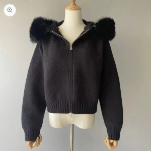 Svart stickad jacka  - Mysig svart stickad jacka med dragkedja och huva med fluffig äkta päls. Jackan har ribbade muddar och midja för en snygg passform. Perfekt för kyliga dagar när du vill vara både varm och stilren.
