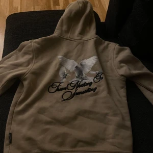 Beige HOODRICH hoodie med duvor - Säljer en beige hoodie från HOODRICH med tryck av vita duvor och texten 'From Nothing To Something' på ryggen. Framsidan har HOODRICH-logga och duvor, samt ficka framtill. Perfekt för dig som gillar streetwear och snygga detaljer.