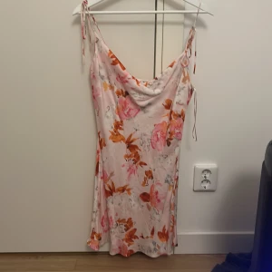 Blommig kortklänning från Zara - Säljer en superfin blommig kortklänning från Zara i ljusa toner med rosa och orange blommor. Klänningen har smala axelband med knyt och en snygg draperad halsringning. Perfekt för varma dagar och sommarhäng!