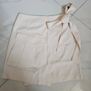 Beige omlottkjol med fickor och knyt - Snygg beige kort omlottkjol med stor ficka framtill och knytband i midjan. Kjolen har dragkedja bak och dekorativa fickor bak. Perfekt för dig som gillar stilrena och enkla plagg med en twist.