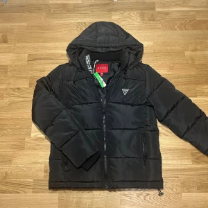 Svart pufferjacka från Guess - Snygg svart pufferjacka från Guess med huva och dragkedja framtill. Jackan har quiltad design, två fickor med dragkedja och diskret Guess-logga på bröstet. Perfekt för kyliga dagar och riktigt skön att bära. Stilsäker och enkel att matcha.
