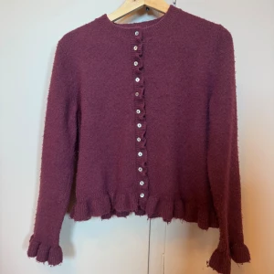 Vinröd kofta med volang från Zara - Vinröd kofta från Zara med volangdetaljer vid ärmslut och nederkant. Tröjan har rund halsringning, knappar framtill och är stickad i ett mjukt material. Den är lite nopprig. Jag säljer den för 200kr + frakt som kunden betalar, priset kan diskuteras.
