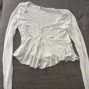 Vit blus med spets från Gina Tricot - Supersöt vit blus från Gina Tricot i storlek XS. Blusen har långa ärmar, volang nertill och spetsdetaljer längs urringningen och en öppning framtill. Med en lätt croppad passform 