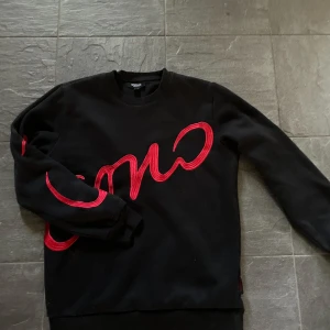 Svart sweatshirt från ICONO med röd text - Svarte sweatshirt från ICONO i regular fit med stor röd broderad text som går över bröstet och ena ärmen. Tröjan har rund halsringning och långa ärmar. Perfekt för dig som gillar streetstyle och vill sticka ut med en cool detalj.