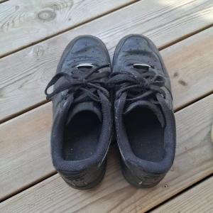 Svarta Nike Air Force 1 sneakers - Klassiska helsvarta Nike Air Force 1 sneakers i skinn med snörning och platt sula. Swoosh-logga på sidorna och perforerade detaljer på tån. Perfekta för dig som gillar en clean och tidlös streetstil.