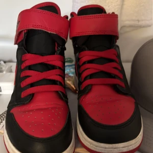 Nike Air Jordan 1 High röd/svart - Säljer ett par Nike Air Jordan 1 High i klassisk röd och svart färg. Skorna har högt skaft, snörning och kardborreband upptill. Tillverkade i skinn med röd skosula. Perfekta för dig som gillar streetstyle.