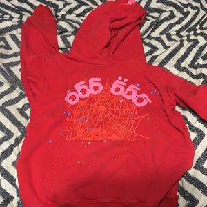 sp5der hoodie - Säljer en röd hoodie med stor huva och magficka. Framsidan har ett coolt tryck med spindelnät i orange och siffror i rosa, samt små blå och vita detaljer. Perfekt för dig som gillar streetwear och unika prints.