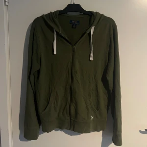 Olivgrön hoodie från Polo Ralph Lauren - Snygg olivgrön hoodie från Polo Ralph Lauren med dragkedja och vita snören i huvan. Tröjan har långa ärmar, två fickor framtill och klassisk logga broderad vid ena fickan. Perfekt för en chill och avslappnad stil.