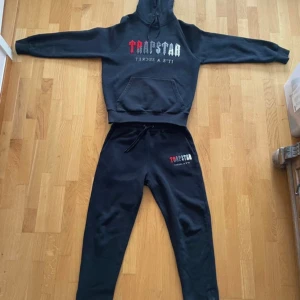 Trapstar svart hoodie med brodyr - Svart hoodie från Trapstar med broderad logga i rött, grått och vitt framtill. Klassisk känguruficka, snörning i huvan och mjukt material. Perfekt för dig som gillar streetwear och vill sticka ut med en fet logga.
