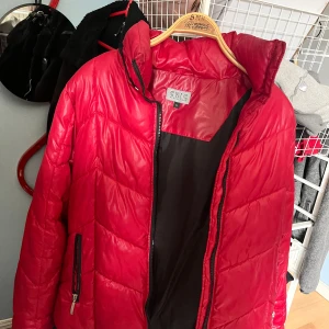 Röd pufferjacka från Chic Casual Wear - Säljer en röd pufferjacka från Chic Casual Wear i storlek M. Jackan har svart innerfoder, dragkedja framtill och en ficka med dragkedja på sidan. Perfekt för kalla dagar och riktigt snygg färg som sticker ut.