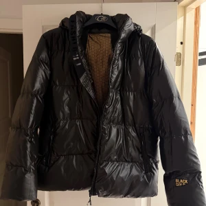 Svart pufferjacka från Fashion Black Gold - Svart pufferjacka från Black Gold med huva och dragkedja framtill. Jackan har quiltad design och en cool detalj med text på ärmen. Insidan har ett mönstrat foder och jackan är perfekt för kalla dagar. Snygg och modern look som passar streetstyle.