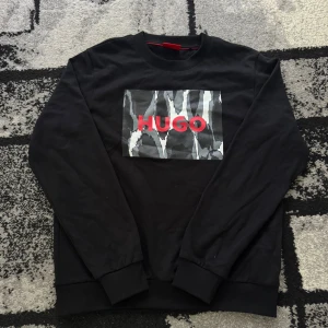 hugo boss tröja - Svart sweatshirt från Hugo med ett stort grafiskt print på bröstet i grått, vitt och svart samt röd HUGO-logga. Tröjan har rund hals, ribbade muddar och är långärmad. Perfekt för dig som gillar streetstyle och vill sticka ut lite extra.