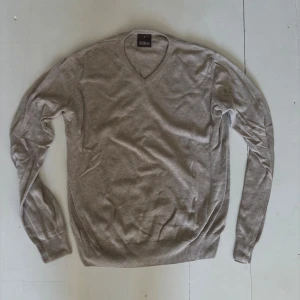 Beige v-ringad tröja Oscar Jacobson - Klassisk beige v-ringad tröja från Oscar Jacobson. Tröjan är i mjuk bomull, har ribbade muddar vid ärmslut och nederkant samt lång ärm. Perfekt för lager-på-lager och enkel att matcha med olika outfits.