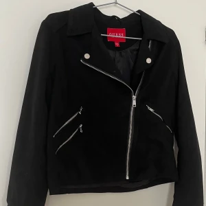 Svart skinnjacka från Guess - Snygg svart skinnjacka från Guess med klassisk krage och flera silvriga dragkedjor framtill. Jackan har lång ärm och en croppad passform som ger en cool vibe. Perfekt för dig som gillar stilrena detaljer och vill ha en jacka som sticker ut.