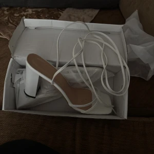 Vita sandaler med snörning och klack - Säljer ett par trendiga vita sandaler med fyrkantig tå och höga blockklackar. Skorna har tunna remmar och långa snören som lindas upp längs benet för en snygg look. Perfekta för att lyfta vilken outfit som helst.