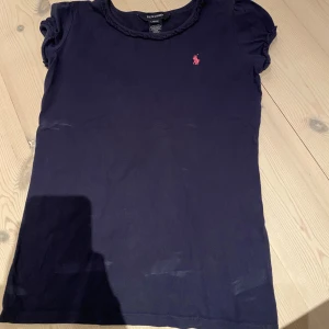 Mörkblå t-shirt från Ralph Lauren - Snygg mörkblå t-shirt från Ralph Lauren med korta ärmar och rosa logobrodyr på bröstet. Rund hals med flätad kant ger en extra detalj. Tillverkad i mjuk 100% bomull för skön känsla hela dagen.