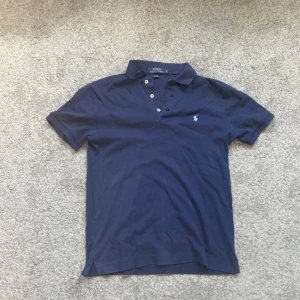 Marinblå Polo Ralph Lauren t-shirt - Snygg marinblå t-shirt från Polo Ralph Lauren i slim fit. Klassisk pikémodell med krage, tre vita knappar och den ikoniska broderade loggan på bröstet. Tillverkad i mjuk bomull som känns skön mot huden. Perfekt för dig som gillar stilrena och tidlösa plagg.