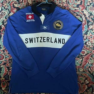 Blå pikétröja Polo Ralph Lauren Switzerland - Blå långärmad pikétröja från Polo Ralph Lauren med svart krage, vit bred rand över bröstet och texten SWITZERLAND. Broderade märken och detaljer framtill, stor romersk siffra IV på ryggen. Skön bomull, perfekt för dig som gillar sportig stil. Säljes på grund av att den har blivit för liten!