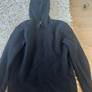Svart hoodie i ull från Oscar Jacobson - Mysig svart hoodie från Oscar Jacobson i mjuk ull. Tröjan har huva och långa ärmar, perfekt för kyliga dagar. Enkel och stilren design utan mönster eller detaljer, passar dig som gillar klassiskt och bekvämt.