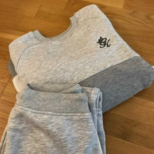 Grå och svart sweatshirt från GK - Säljer två snygga sweatshirts från GK, en i ljusgrått med mörkare grå panel och svart broderad logga, och en i helsvart med guldfärgad broderad logga. Båda har rund hals och ribbade muddar. Perfekt för chill dagar eller streetstyle.( Båda för 300 1 för 180)