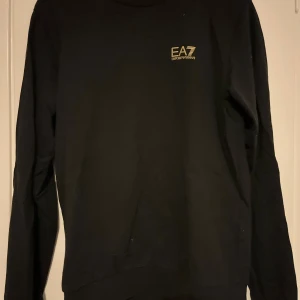 Svart EA7 långärmad tröja - Svart långärmad tröja från EA7 Emporio Armani med diskret guldfärgad logga på bröstet. Enkel och stilren design, perfekt för dig som gillar klassiska streetwear-vibbar. Mjuk och bekväm med rund halsringning.
