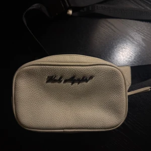 Beige väska från Karl Lagerfeld - Snygg beige väska från Karl Lagerfeld i grynigt skinn. Väskan  har svart justerbart band med logotyp och smidig plastspänne. Perfekt för att bära det viktigaste och ge din outfit en clean vibe.