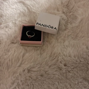 Pandora silverring med röd hjärtformad sten - Superfin silverring från Pandora med en röd hjärtformad sten i mitten och små detaljer runt hela ringen. Perfekt för dig som vill ha något gulligt och romantiskt på fingret. Kommer i originalask. 