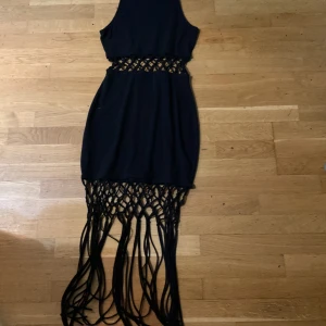 Svart klänning med fransdetaljer - Svart ärmlös klänning med coola utskurna detaljer i midjan och långa fransar nertill. Klänningen har en tight passform upptill och ett unikt nätmönster som ger en edgy vibe. Perfekt för dig som vill sticka ut med en statement piece. Köpt för 499 säljer för 200