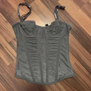 Grå mesh korsett från H&M Divided - Säljer en grå korsett-topp från H&M Divided i mesh med justerbara axelband och snygga rynkade detaljer framtill. Toppen har byglar vid bysten och hakknäppning bak för en tight passform. Perfekt för att lyfta vilken outfit som helst.