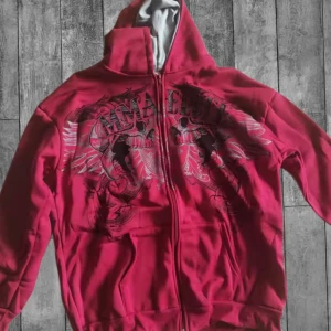 Unik vintage zip hoodie - Säljer en röd vintage hoodie med stort tryck av dödskallar och vingar på framsidan. Tröjan har dragkedja, huva och två fickor framtill. Perfekt för dig som gillar streetwear och vill sticka ut med en cool design.