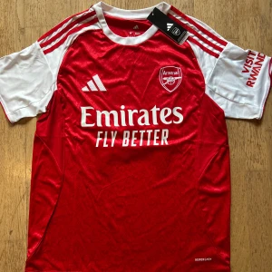 Arsenal Gyökeres 14 matchtröja Adidas - Arsenal fotbollströja med Gyökeres och nummer 14 på ryggen. Röd kropp med vita ärmar, klassiska Adidas-ränder på axlarna och klubbmärke på bröstet. Tillverkad i lätt och ventilerande material, perfekt för match eller träning. Skriv privat för mer frågor. 