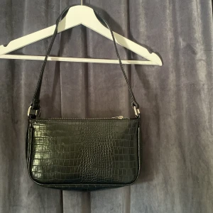 Svart axelväska i croco-imitation - Snygg svart axelväska med croco-mönstrad yta och silvrig dragkedja. Väskan har en stilren, rektangulär form och ett justerbart axelband. Perfekt storlek för det viktigaste och ger en trendig touch till din outfit.