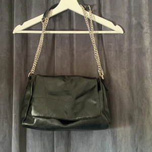 Svart axelväska med kedjerem från vero Moda  - Snygg svart axelväska med mjukt material och silverfärgad kedjerem. Väskan har lock framtill, innerfack med dragkedja och stilrena sömdetaljer. Perfekt storlek för att få plats med allt du behöver och passar till många olika outfits.