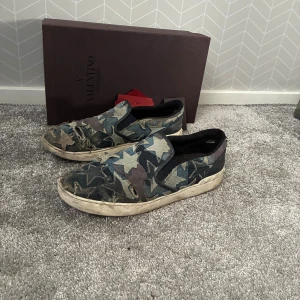 Valentino Garavani slip-on sneakers - Slip-on sneakers från Valentino Garavani med ett unikt camouflage-mönster i blått, grönt och beige. Vill även tillägga att skorna inte är rengjorde och tvättade på länge vilken gör att de ser extremt smutsiga ut. De är dock använda en hel del och har små defekter på sulan under men de påverkar inte användningen (: Dessa skorna är extremt sällsynta och är defensivt svåra att lägga sina händer på✌🏼