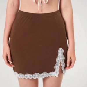 Brun kortkjol med vit spets - Supersöt brun kortkjol med elastisk midja och vit spetsdetalj längs nederkanten och en snygg slits framtill. Kjolen har en enkel, rak form och är perfekt för dig som gillar en feminin och trendig look. Materialet känns mjukt och bekvämt mot huden.